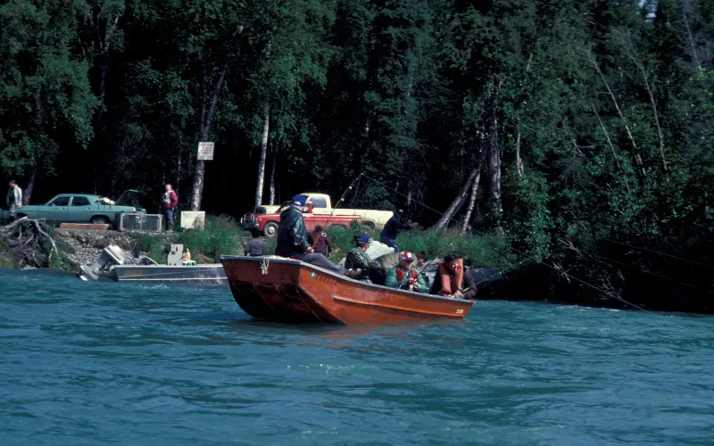 A boat operator stands at the stern while anglers in life jackets fish from a riverboat on the Kenai River in Alaska.