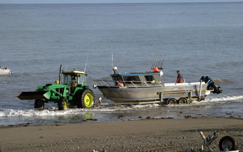 A tractor tows an aluminum charter fishing boat on a trailer through the surf at Anchor Point, Alaska, as a deckhand stands on deck by the rod rack