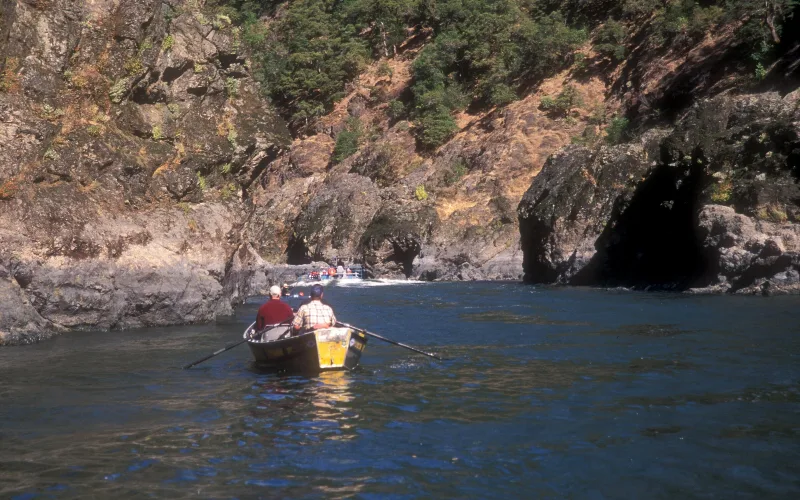 Two men in a yellow drift boat row down the Rogue River in southern Oregon as a jet boat approaches through a rocky canyon.