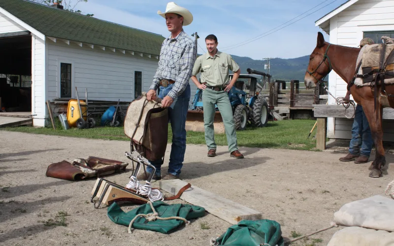 A packer in a cowboy hat holds a pack saddle over canvas packing tarps during a backcountry packing demonstration at the Ninemile Ranger District, Lolo National Forest, with a loaded mule and a Forest Service employee nearby.