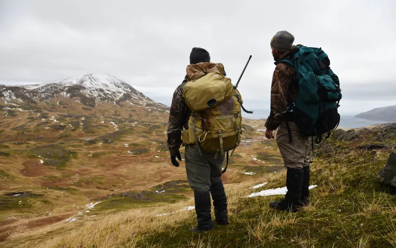 Two hunters with backpacks and a rifle stand on a grassy ridge looking out over tundra and a snow-dusted peak on Adak Island, Alaska.