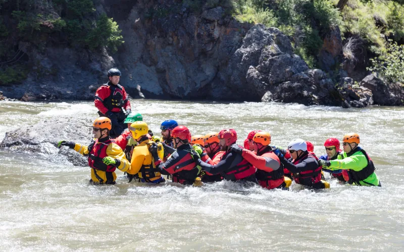 Students in helmets, drysuits and life jackets link arms in a wedge to wade across a fast river during a swiftwater rescue training course in Yellowstone National Park