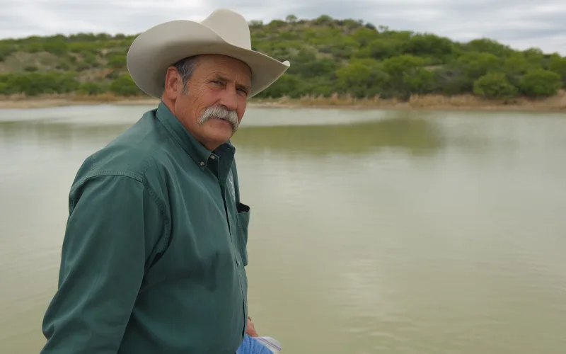 A ranch manager in a cowboy hat and green work shirt beside a stock pond on a Texas cattle ranch, with brushy hills behind.