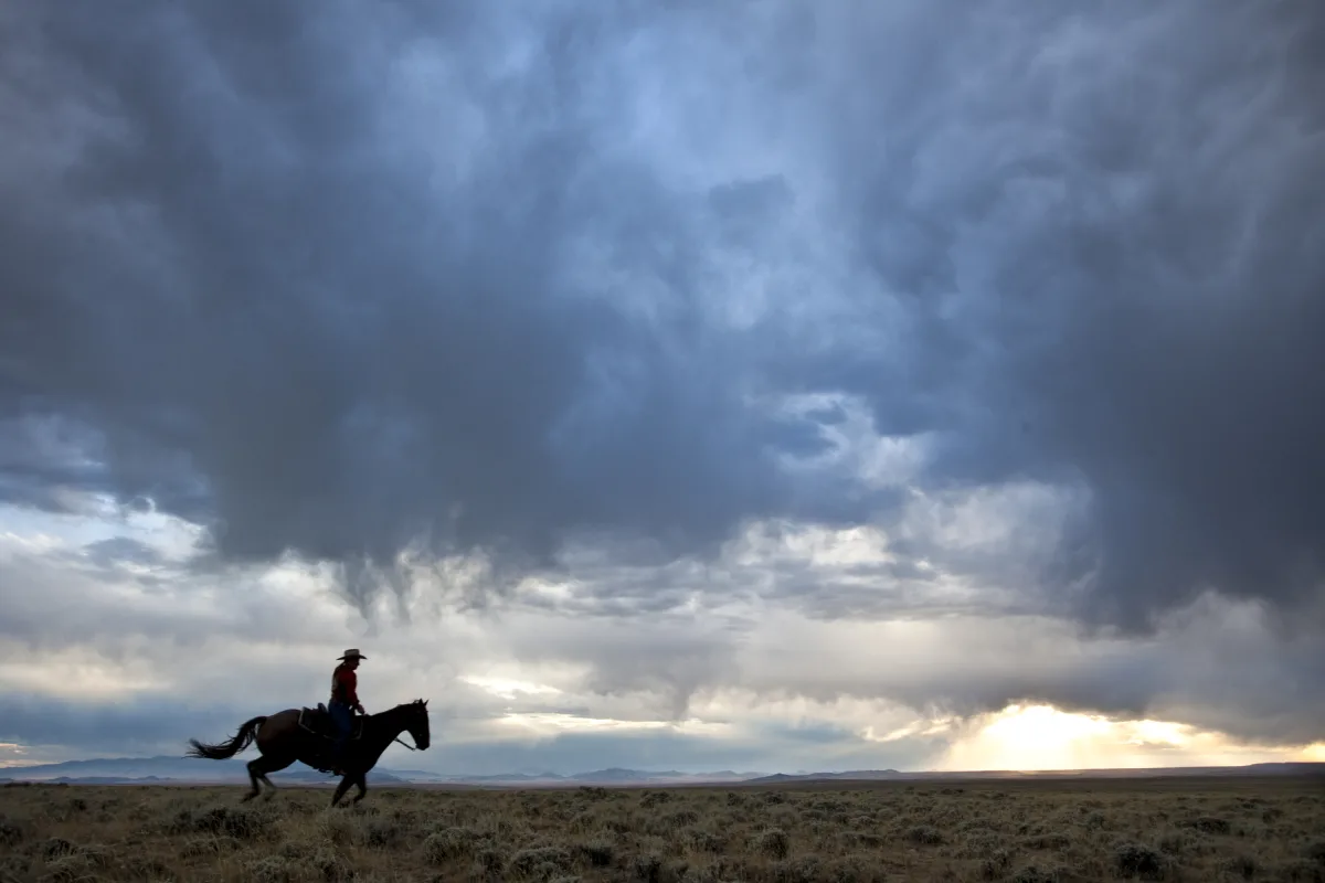 A rider galloping across open sagebrush country under a dark storm sky with late light breaking on the horizon.