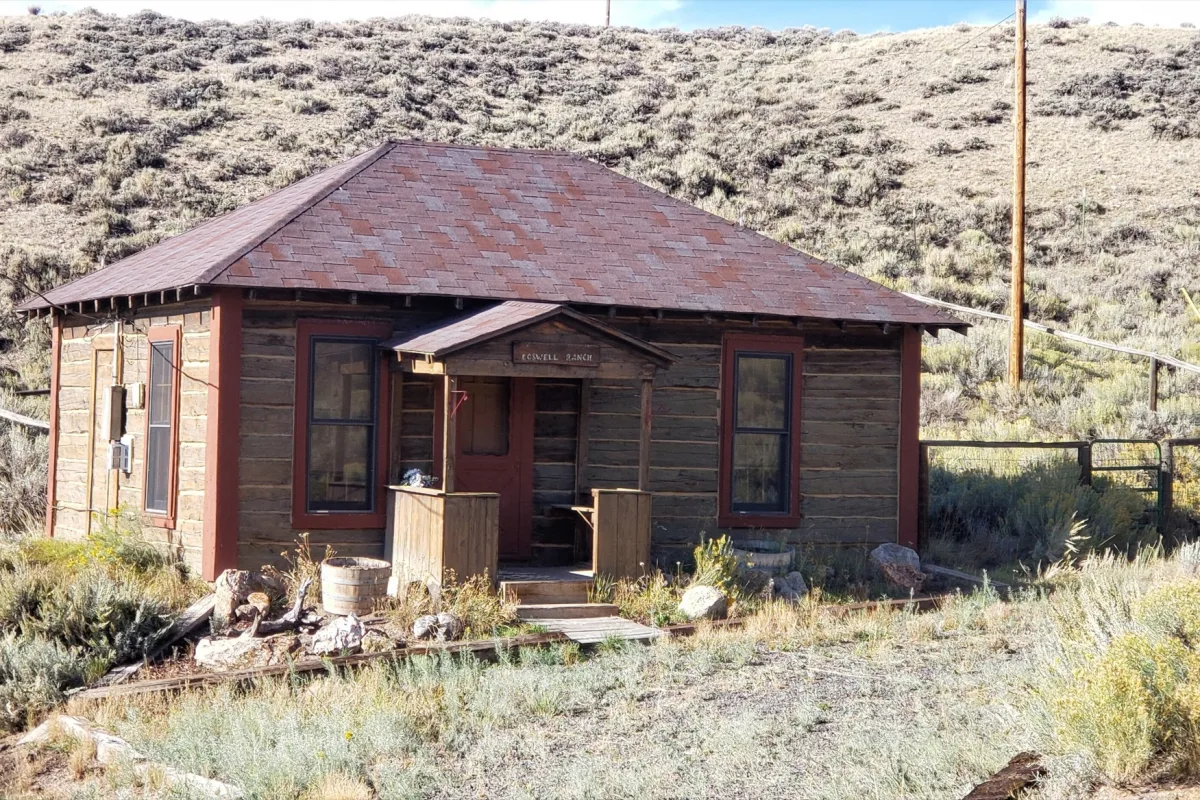 A weathered wooden ranch bunkhouse with red trim and a hip roof sits among sagebrush below a dry hillside in Albany County, Wyoming.