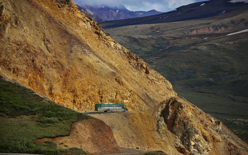 A green park bus drives a gravel road cut into the side of Polychrome Mountain in Denali National Park, with tundra valleys and peaks behind