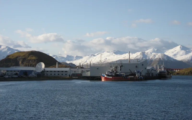 Seafood processing plants and a red fishing boat at the dock in Dutch Harbor, Alaska, with snow-covered mountains behind.