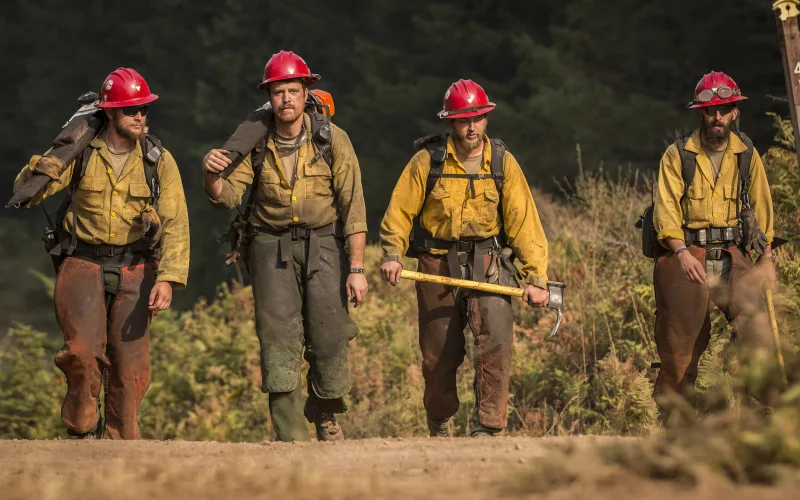 Four hand crew firefighters in yellow shirts, red helmets and leather chaps walk up a dirt road carrying saws and a Pulaski.