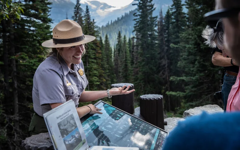 A National Park Service ranger in a flat hat and gray uniform points to an exhibit panel while talking with visitors at Jackson Glacier Overlook, with forest and mountains behind her