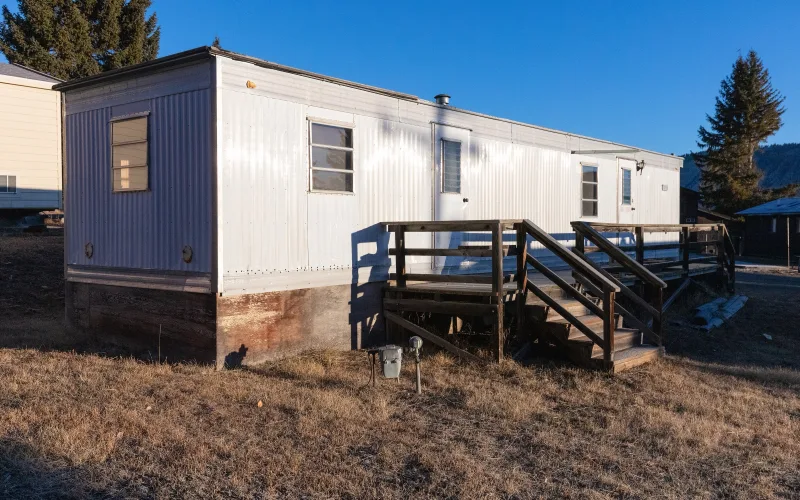 A white single-wide trailer used as seasonal employee housing in Yellowstone National Park, with a wooden deck and stairs, dry grass and pine trees under a blue sky.